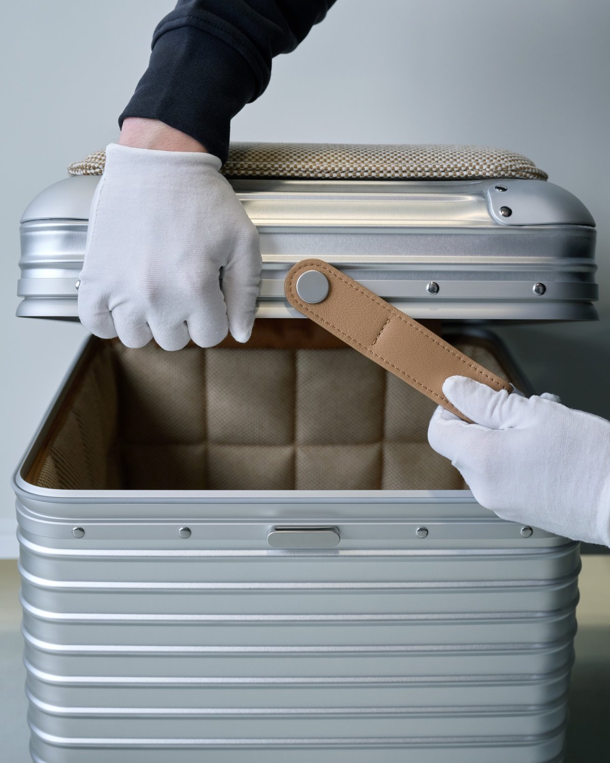 Two hands wearing white gloves lift the lid of a grooved aluminum Rimowa Vitra stool.