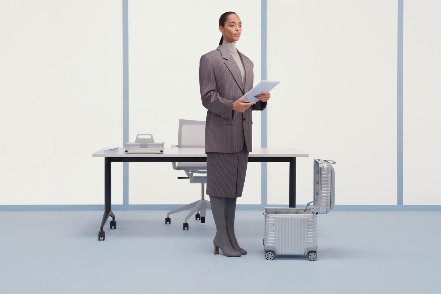 A woman in a grey suit stands next to an aluminum Rimowa Vitra stool, with a matching grooved aluminum toolbox on the desk behind her.