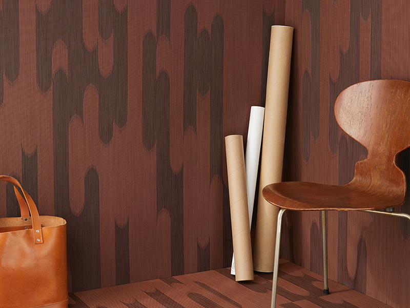 Brown and brick red Shuttle carpeting by Chilewich with a wooden chair, rolled paper and leather bag in the foreground