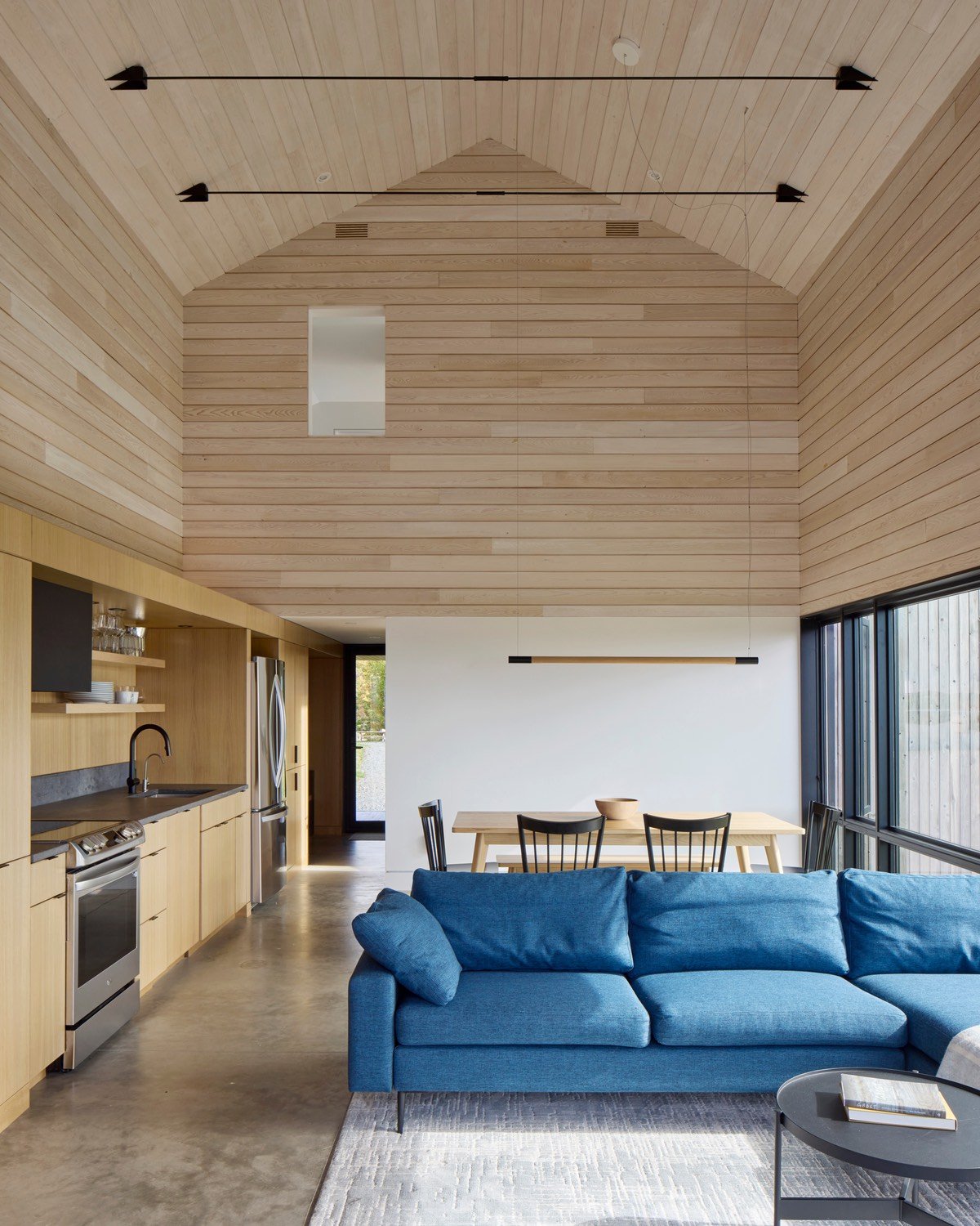 A blue sofa sits in front of a wooden dining table with a kitchen to the left inside a wooden cabin with a pitched roof.