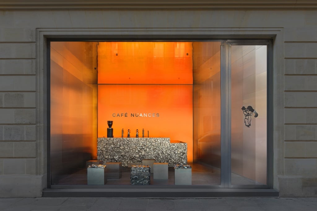 A view from the street into Cafe Nuances in the Marais neighbourhood of Paris designed by Crosby Studios. Four cube stools sit in front of a textural counter clad in cast steel versions of crumpled coffee cups.