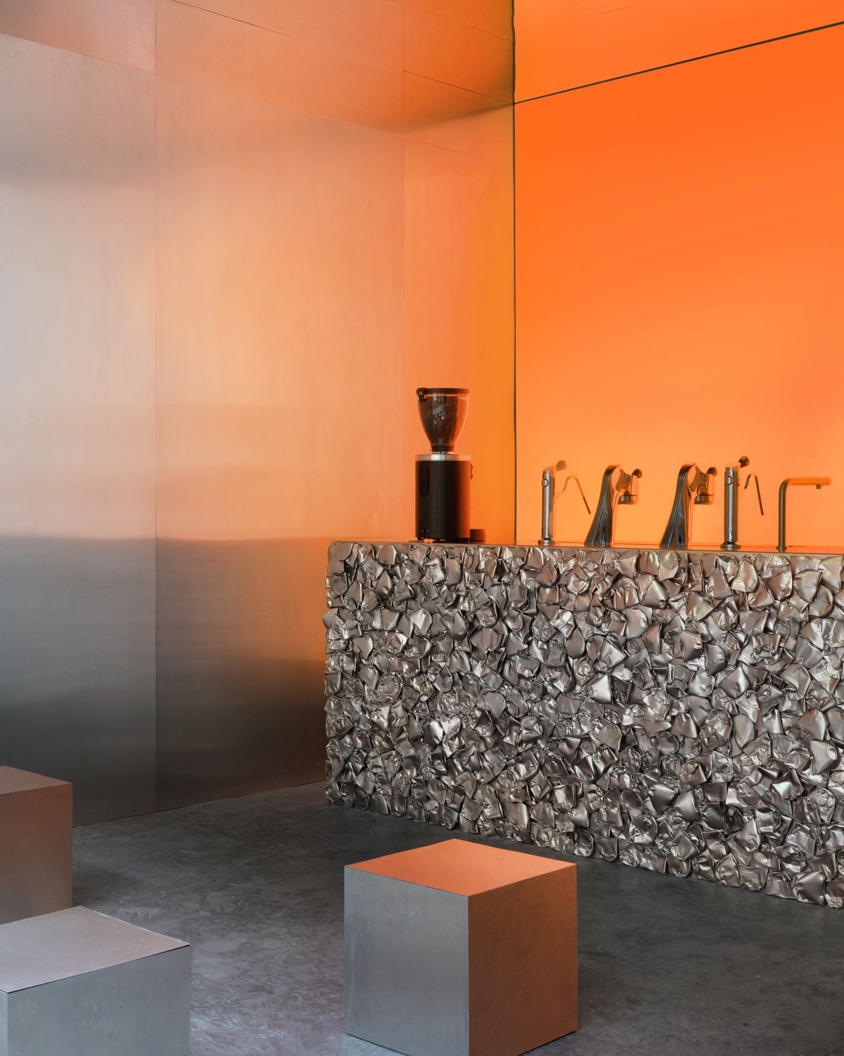 A stainless steel bar decorated with steel casts of crumpled coffee cups is installed in front of a glowing orange wall at Café Nuances in the Marais neighbourhood of Paris designed by Crosby Studios..