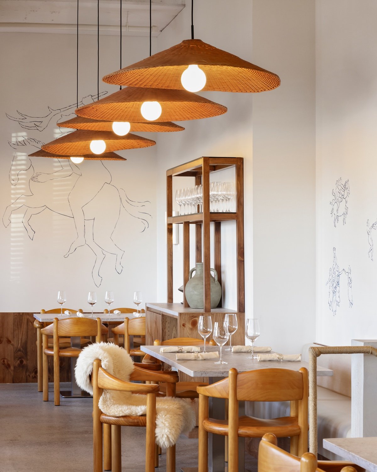 Rattan lampshades hang above a series of curved pinewood chairs inside Selene, a Vancouver Greek restaurant by design studio &Daughters.