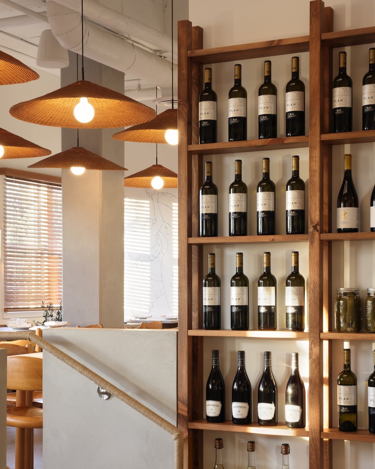 Pine shelves hold rows of wine bottles. A rope-wrapped railing leads up to an elevated dining area lit by hanging rattan lampshades inside Selene, a Vancouver Greek restaurant by design studio &Daughters.