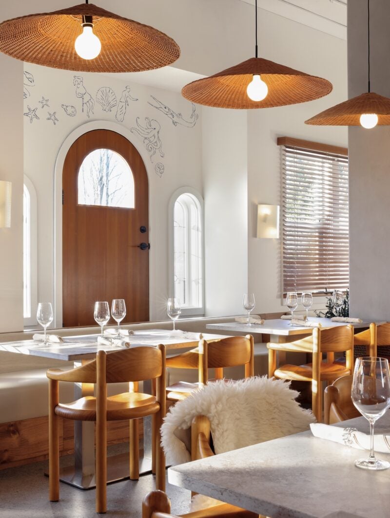 Rattan lampshades hang above a row of curved pinewood chairs seated at dining tables in Vancouver Greek restaurant Selene designed by &Daughters.