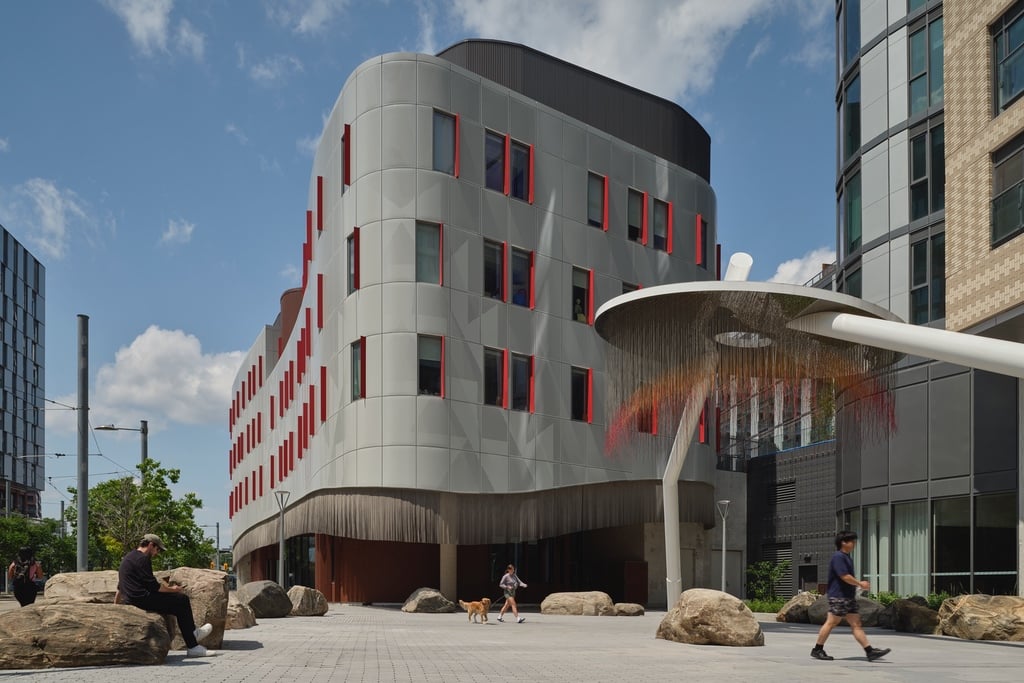 The Toronto Indigenous Hub is anchored by a curving building with a border of stainless steel beads hanging over the entrance. To the right is a public artwork that is a floating disc with red and orange strings hanging down from it.