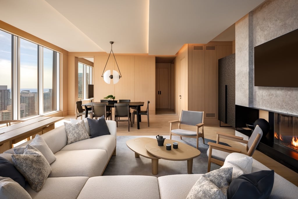 A living room zone in one of the guest rooms at Nobu Hotel Toronto featuring a plush beige sofa that looks towards a TV mounted on a grey marble wall. In the back is a six-person dining table with a circular pendant light hanging above.