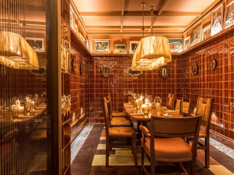 A private dining room clad in burgundy red tiles features a row of sculptural plates featuring seafood dishes and above it, a row of still life paintings.