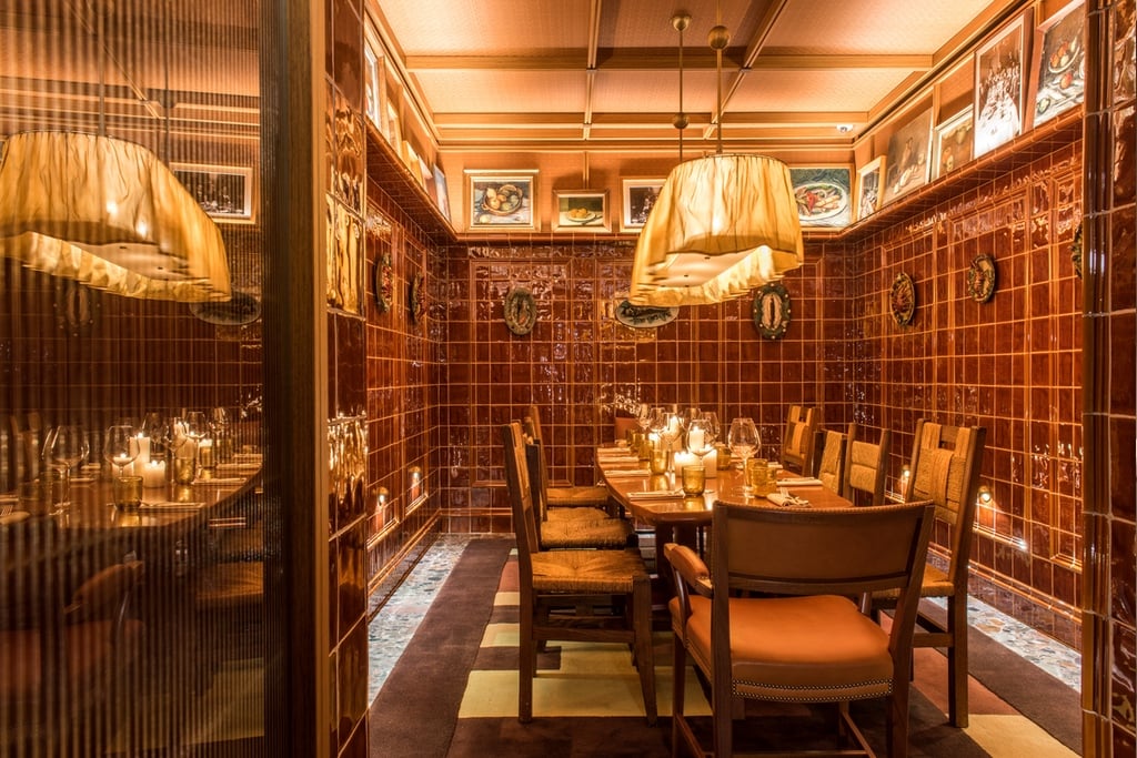 A private dining room clad in burgundy red tiles features a row of sculptural plates featuring seafood dishes and above it, a row of still life paintings.