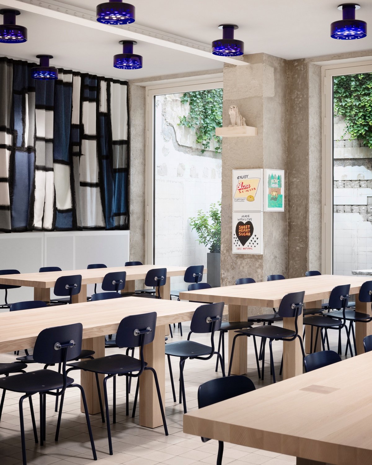 A room full of inky blue chairs seated at blonde wood dining tables in front of a curtain with a grid of blue, white and grey squares. The ceiling features purple glass sconces.
