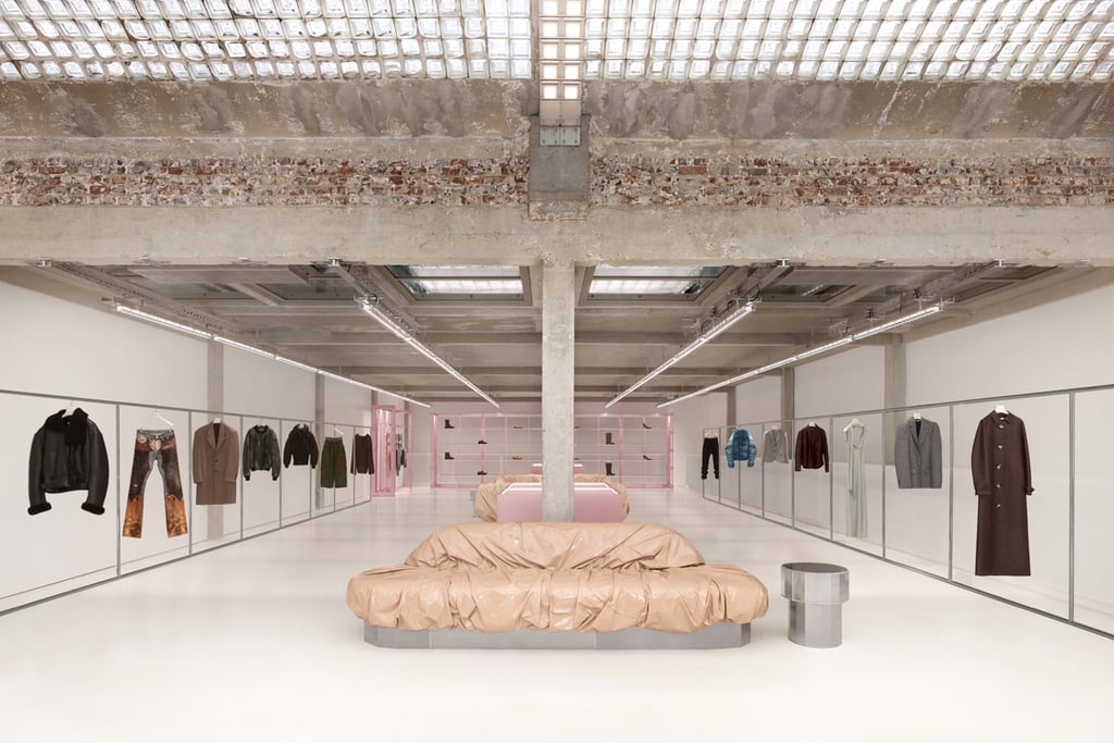 A glass-brick ceiling illuminates the Paris showroom of Acne Studios, featuring clothing racks on either side of the concrete room and a sofa wrapped in crumpled, cream-coloured vinyl at the centre. It was designed by Halleroed.