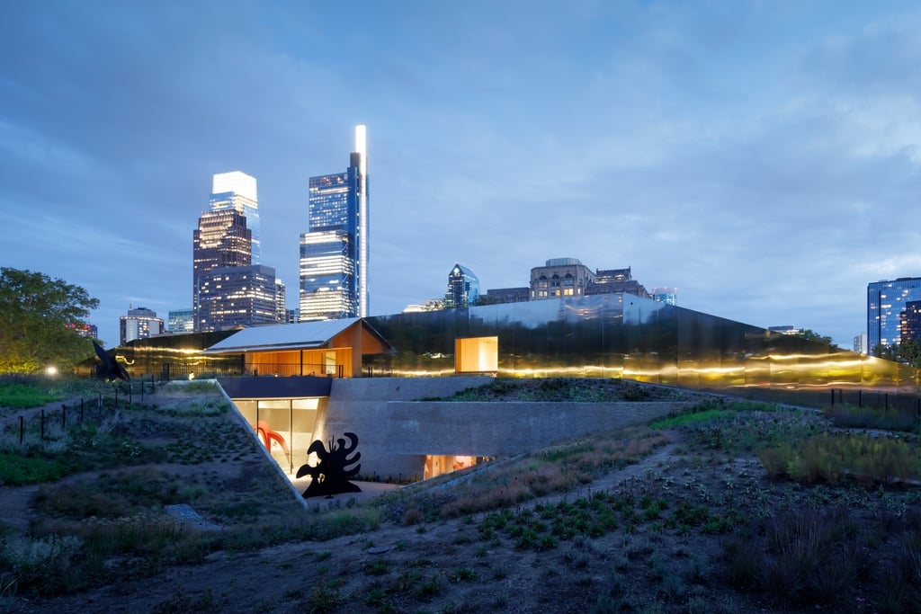 A view of Calder Gardens at night, showing artworks in a sunken garden gallery with the city skyline in the background.