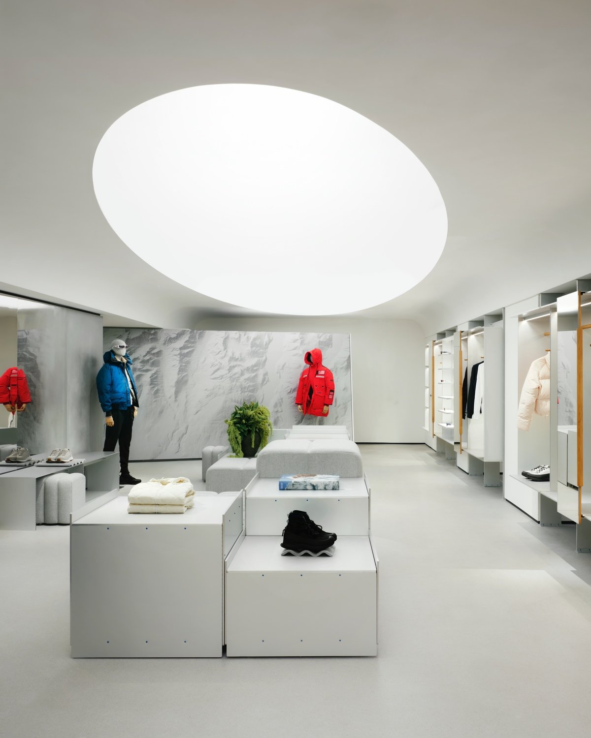 The Canada Goose store in Paris designed by Snohetta featuring a glowing oculus in the ceiling above racks of jackets and square aluminum display podiums with winter boots on them.
