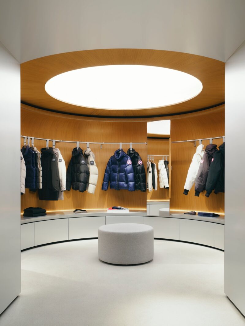 The Canada Goose store in Paris designed by Snohetta featuring a glowing oculus in a rounded room with a clothing rack showcasing a row of parkas and puffer jackets.