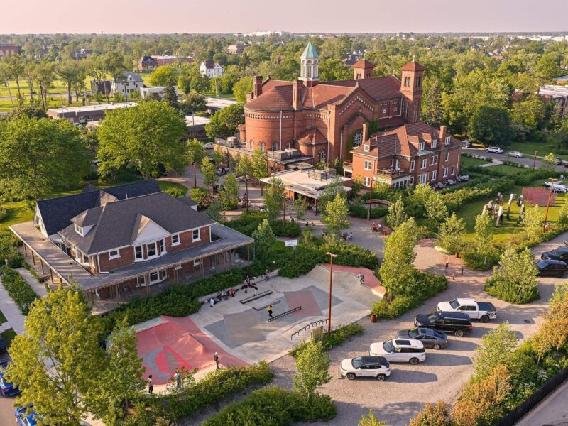 Aerial view of a skate park, parked cars, and two large brick buildings in Detroit surrounded by trees on a sunny day.