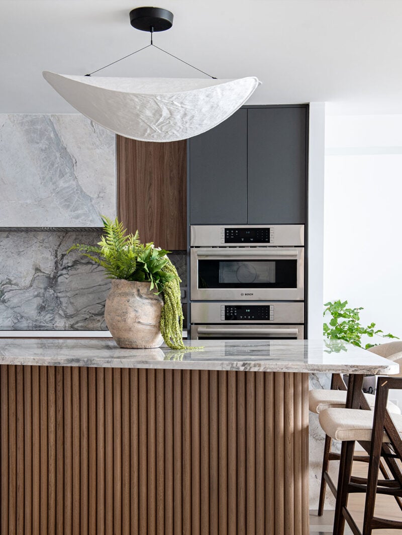 A stack of ovens from Bosch Appliances are shown in a Toronto kitchen with a curving island topped in marble and clad in large wooden dowels. Above it is a paper pendant light.