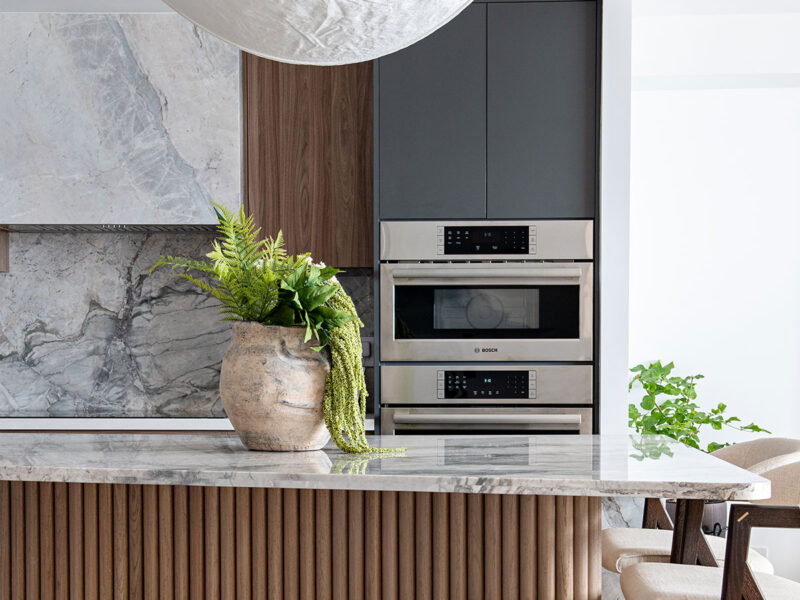 A stack of ovens from Bosch Appliances are shown in a Toronto kitchen with a curving island topped in marble and clad in large wooden dowels. Above it is a paper pendant light.