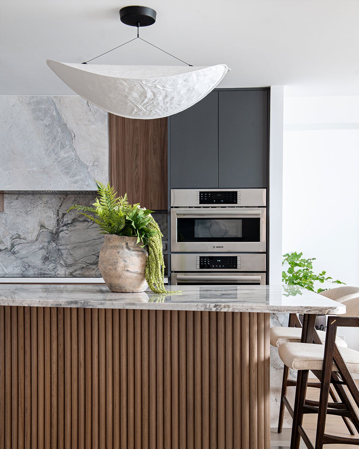 A stack of ovens from Bosch Appliances are shown in a Toronto kitchen with a curving island topped in marble and clad in large wooden dowels. Above it is a paper pendant light.
