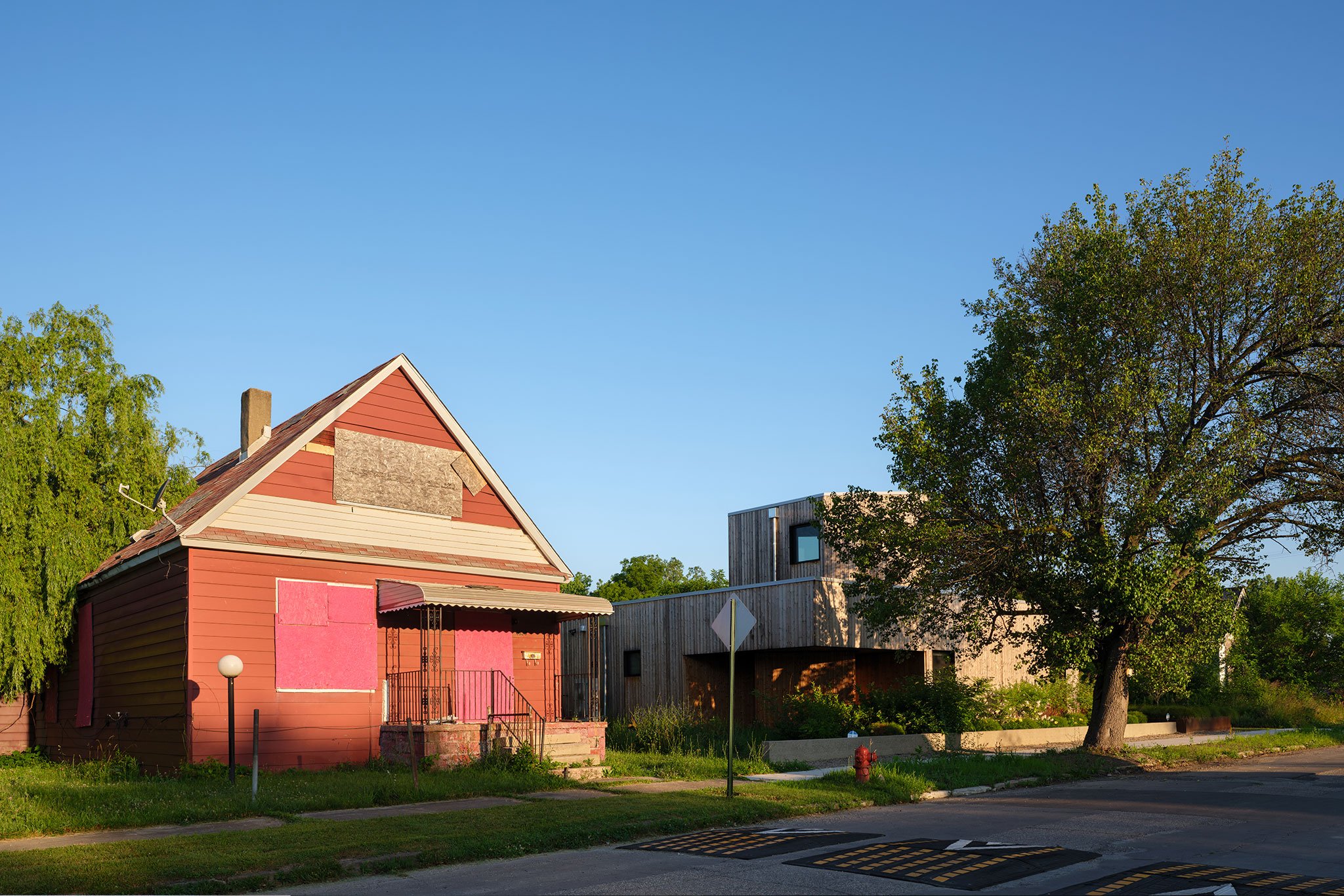 A view from the street of an old boarded-up red house next to a new modern home clad in wood.