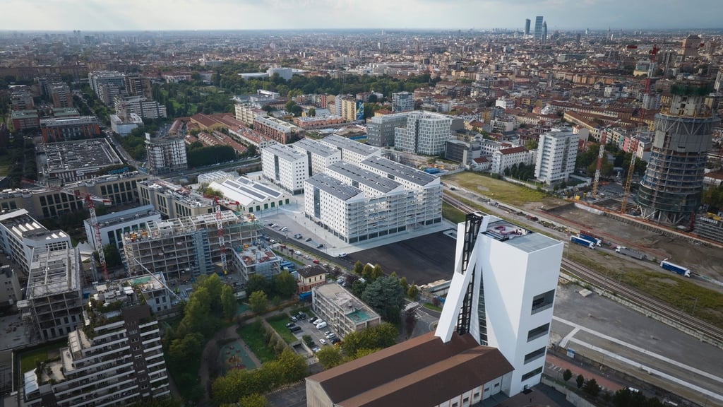 An aerial view of the athlete's village at the 2026 Milan Cortina Olympics, showing a cluster of white residential mid-rise buildings.