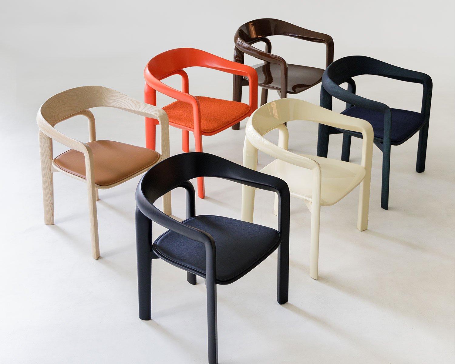 Two rows of chairs with curved backrests shown in a variety of colours, including a glossy red and a natural wood with a brown leather cushion. It's a preview of a chair being showcased at IDS Toronto 2026.