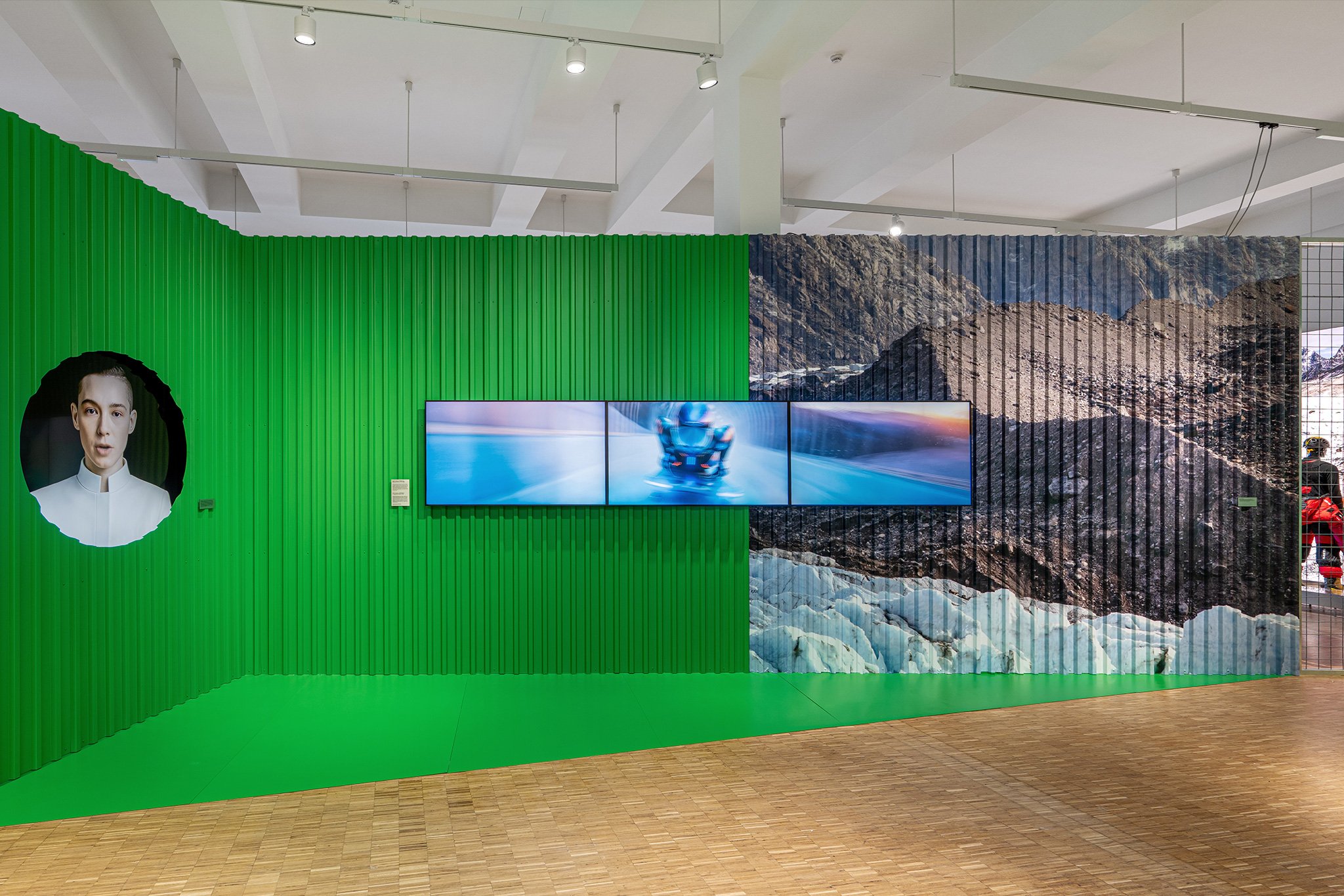 A corrugated metal wall inside a gallery at the Triennale design museum. Half of the wall is green and half of the wall features an image of a ski chalet. At the centre are three screens playing futuristic animations.
