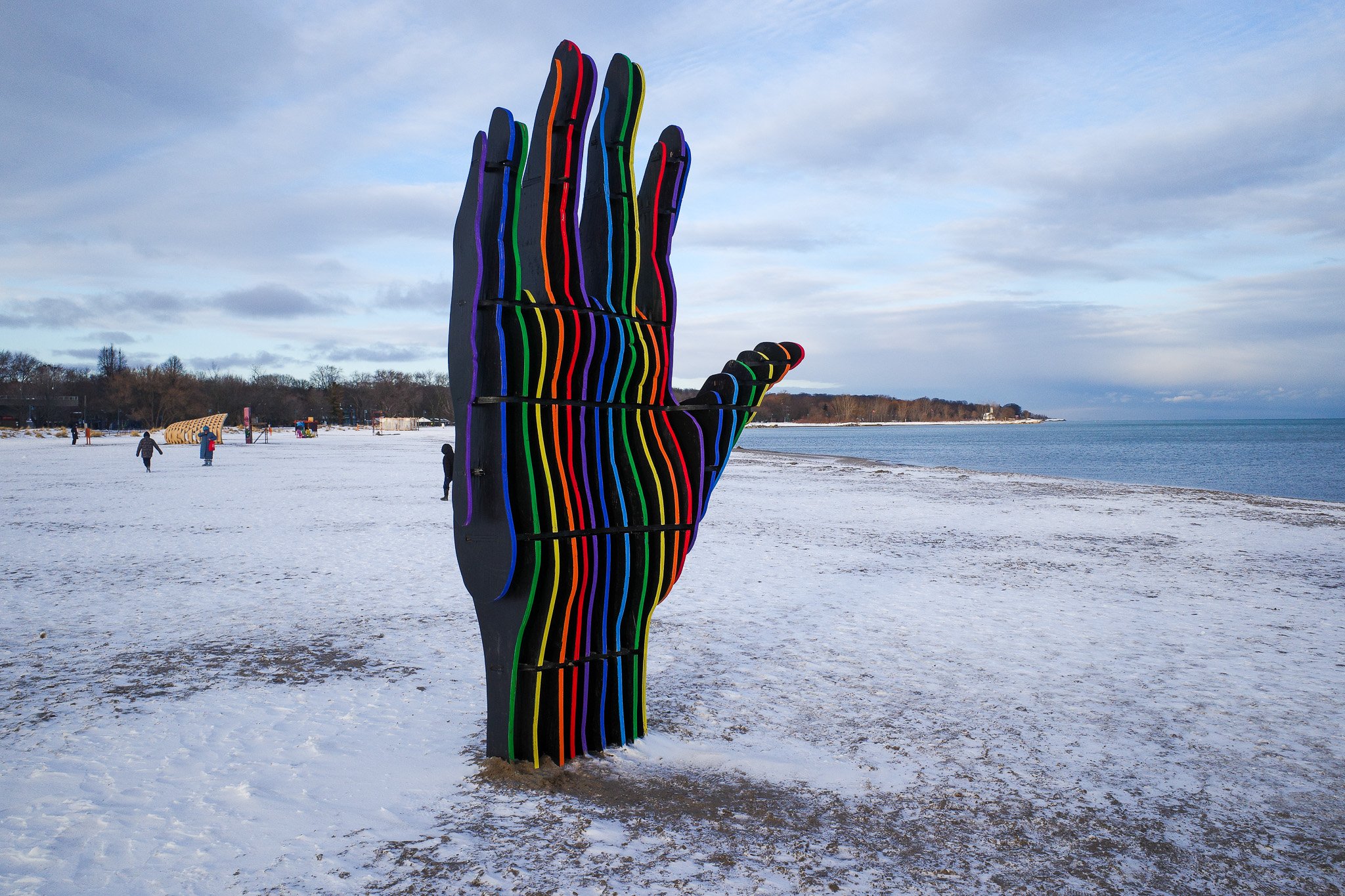 A wooden sculpture of a hand installed on a snow-covered beach and built from slices of wood that are each painted a different colour.