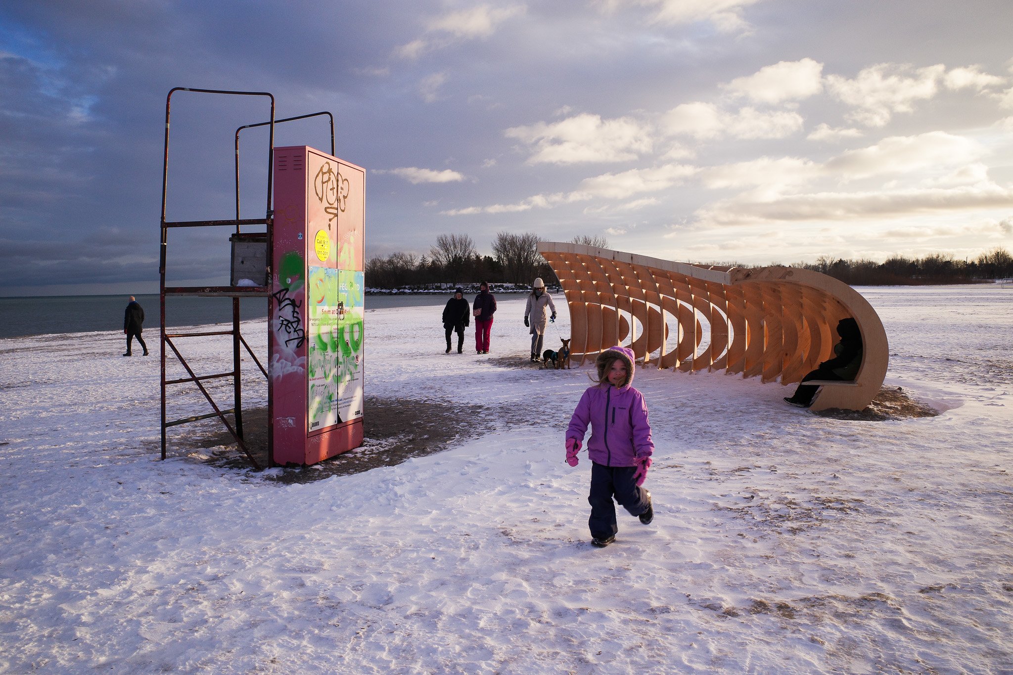 A child runs in front of a wave-shaped wooden structure installed on a snowy beach next to a lifeguard stand as part of Toronto's Winter Stations 2026 exhibition.