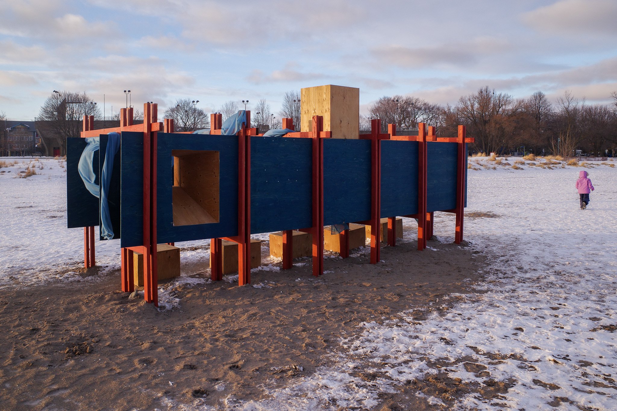 A long, rectangular wood-framed structure painted blue and red with a window at the end installed along the snowy beach as part of Toronto's Winter Stations 2026 festival.