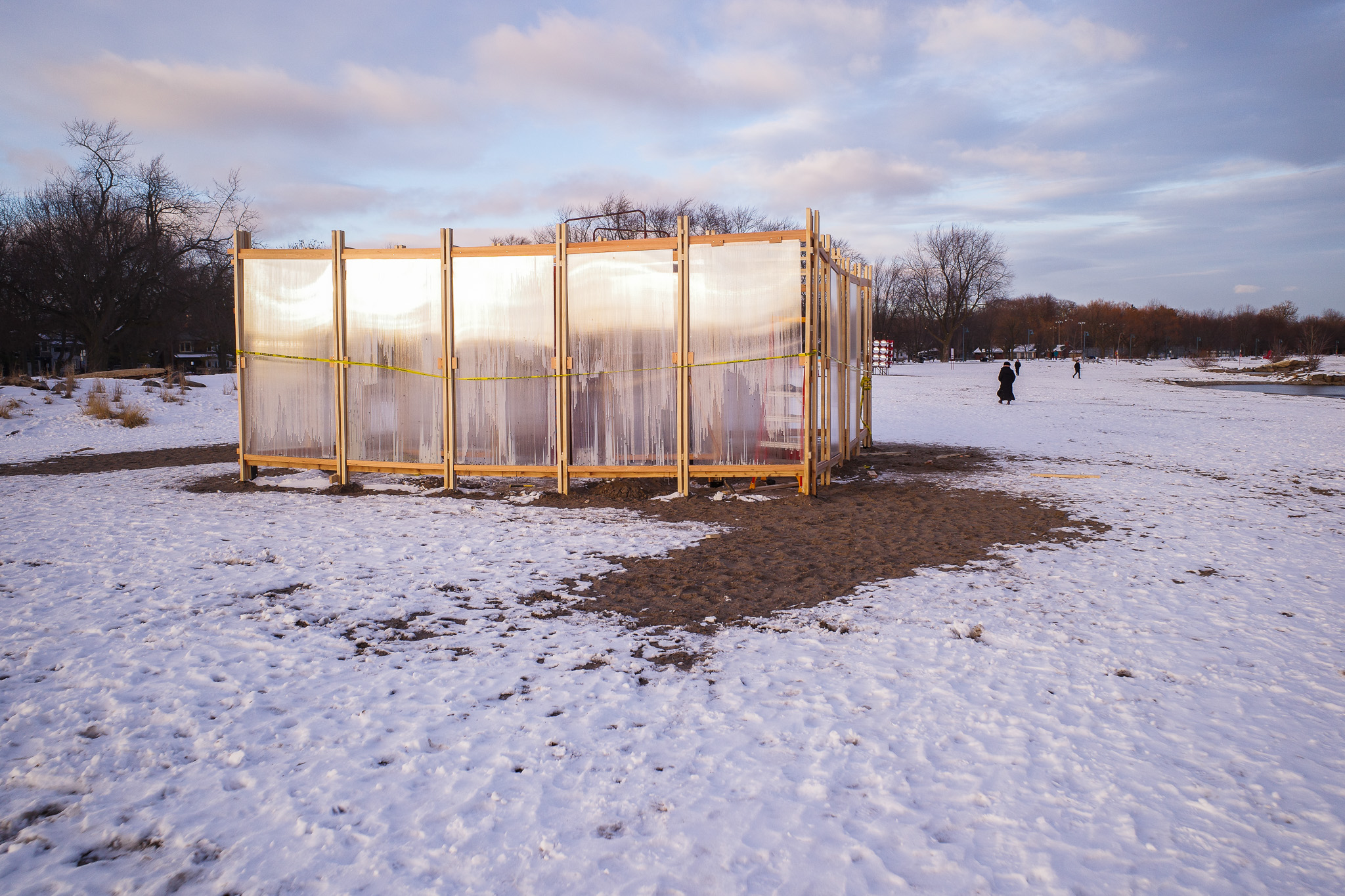 A person walks along a snow-covered beach past a plywood-framed pavilion with translucent polycarbonate panels at Winter Stations 2026 in Toronto.