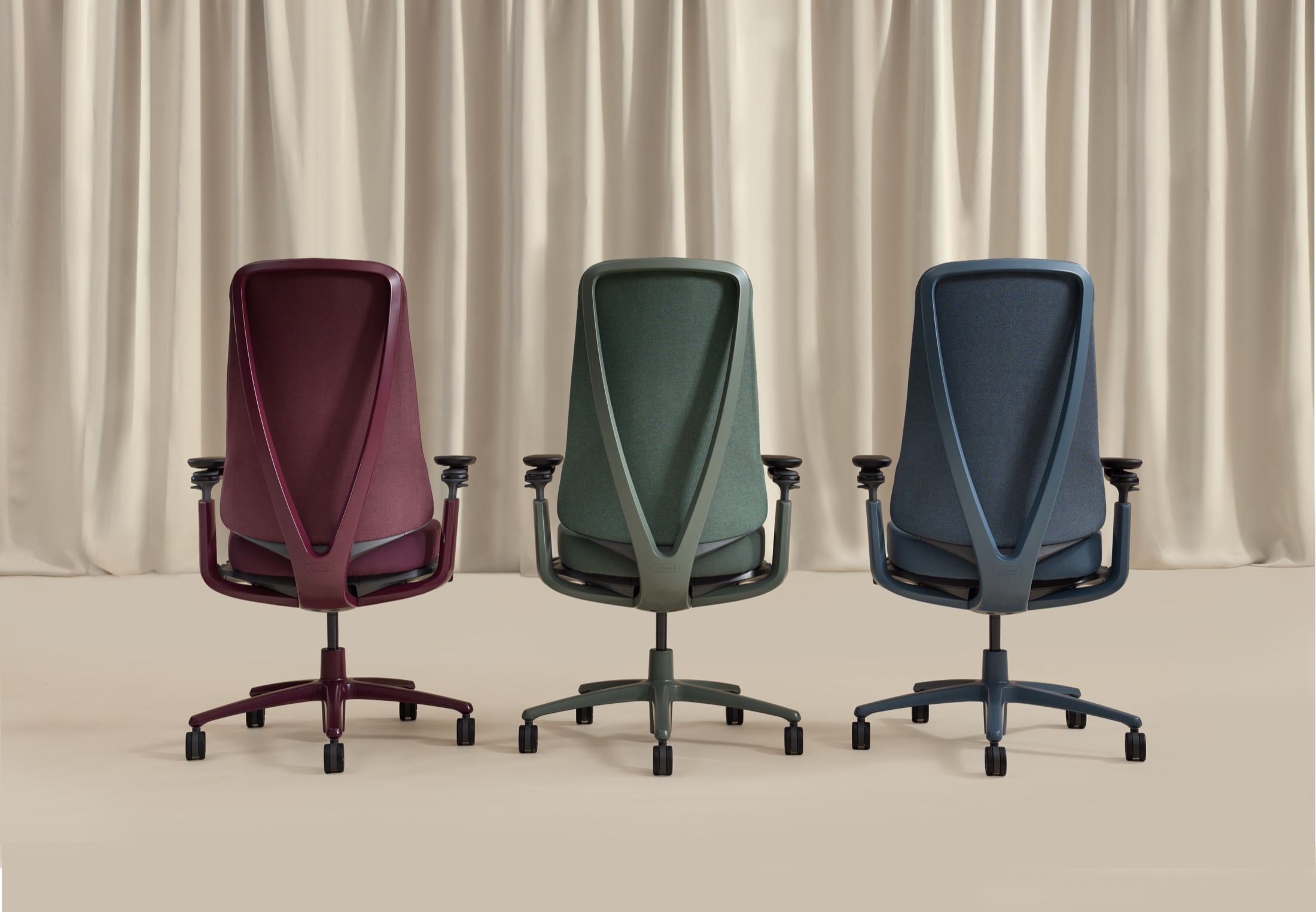 Three ergonomic office chairs in burgundy, green, and blue are shown from the back, positioned side by side in front of beige curtains.
