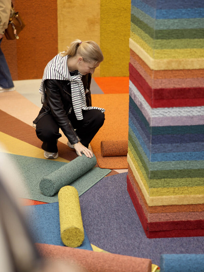 A person kneels on a colorful carpet floor, rolling up a green textured rug beside a tall stack of multicolored rolled carpets at Copenhagen Design Week 2026.