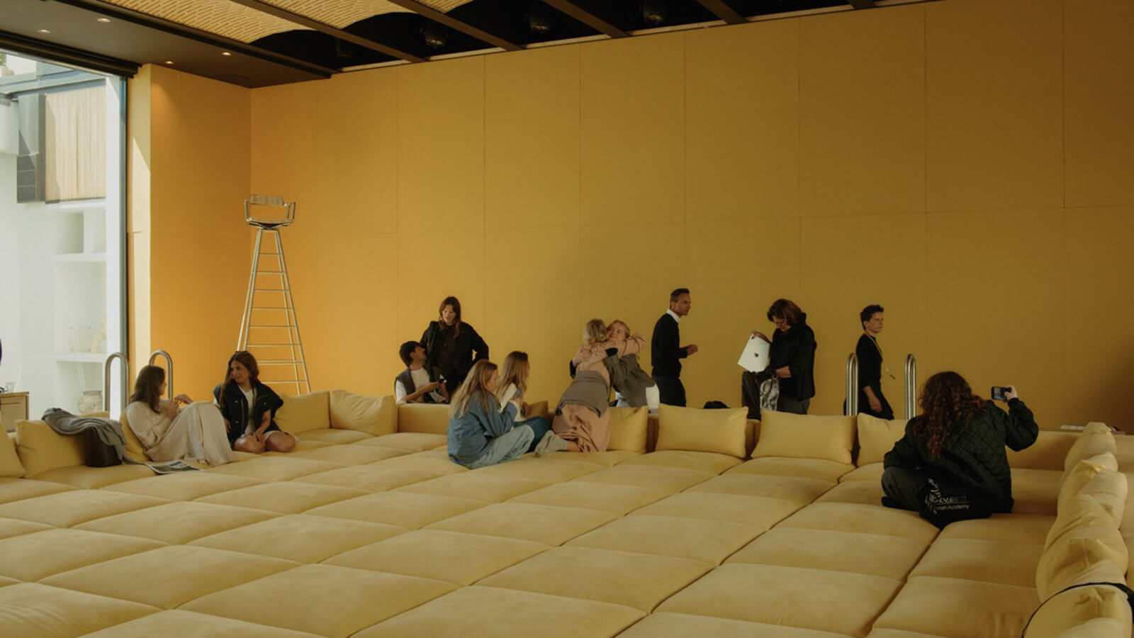 A group of people sit and stand on a large, yellow, cushioned platform in a minimalist room with yellow walls and a ladder in the background at Copenhagen Design Week 2026.