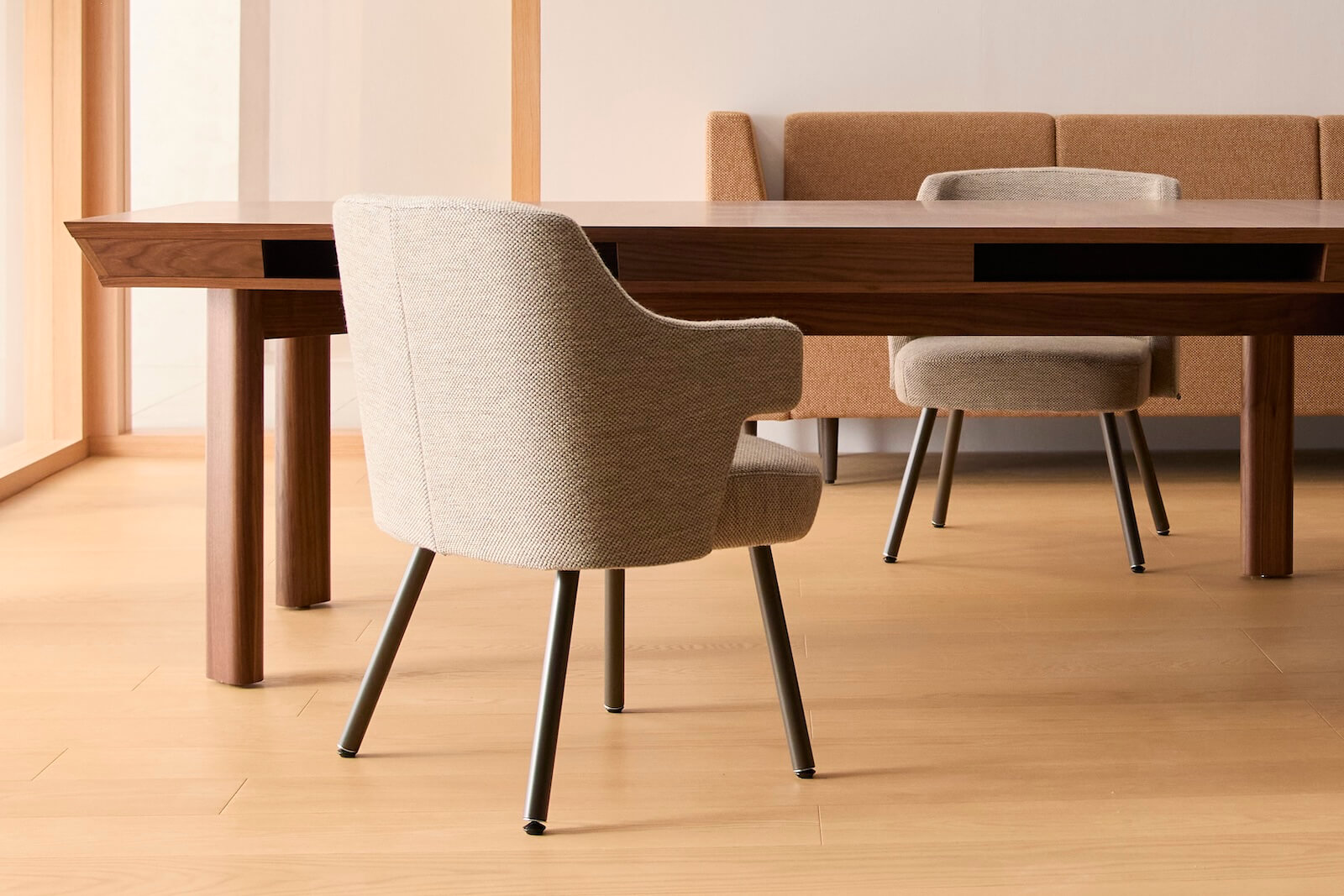 A beige upholstered armchair with black legs is placed near a wooden table; another matching chair is partially visible in the background on a light wood floor.