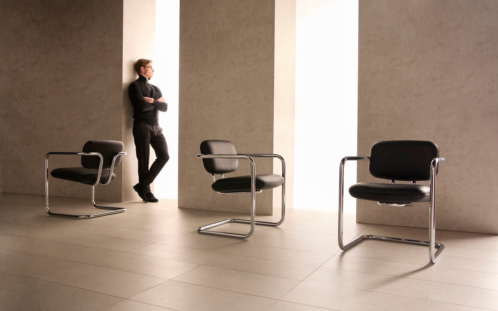 A man in black clothing stands against a wall by a tall window; two empty black chairs with metal frames are in the foreground on a tiled floor.