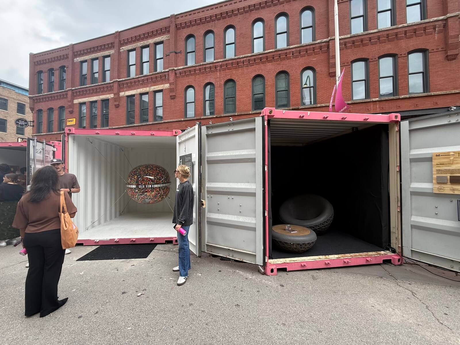 Three people stand near two open shipping containers displaying art installations on a city street at Fulton Market DesignDays 2026, with a brick building in the background.