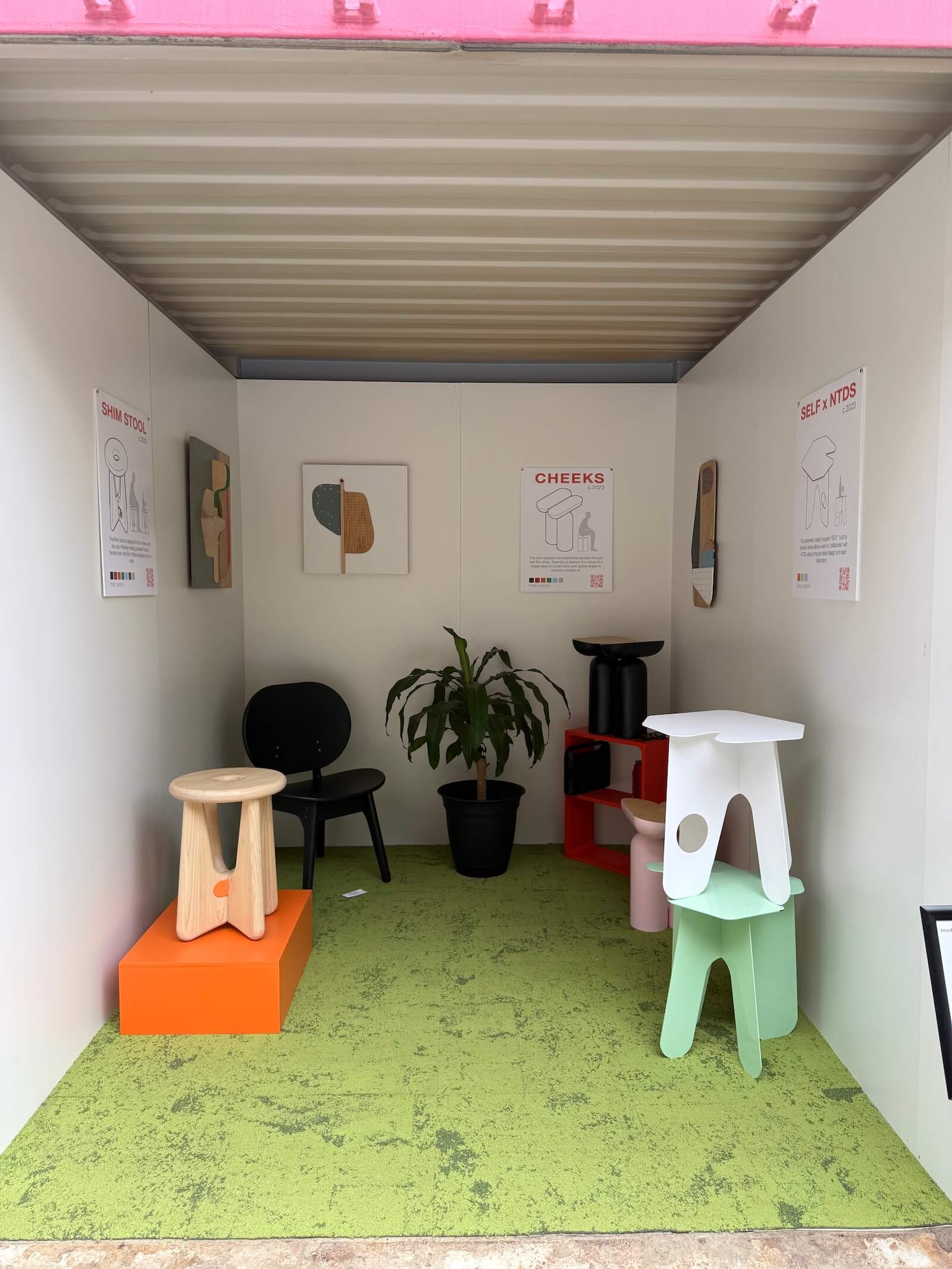 A small exhibition space at Fulton Market DesignDays 2026 displays modern stools, a potted plant, and wall art on white walls with green flooring and posters showing product names and illustrations.