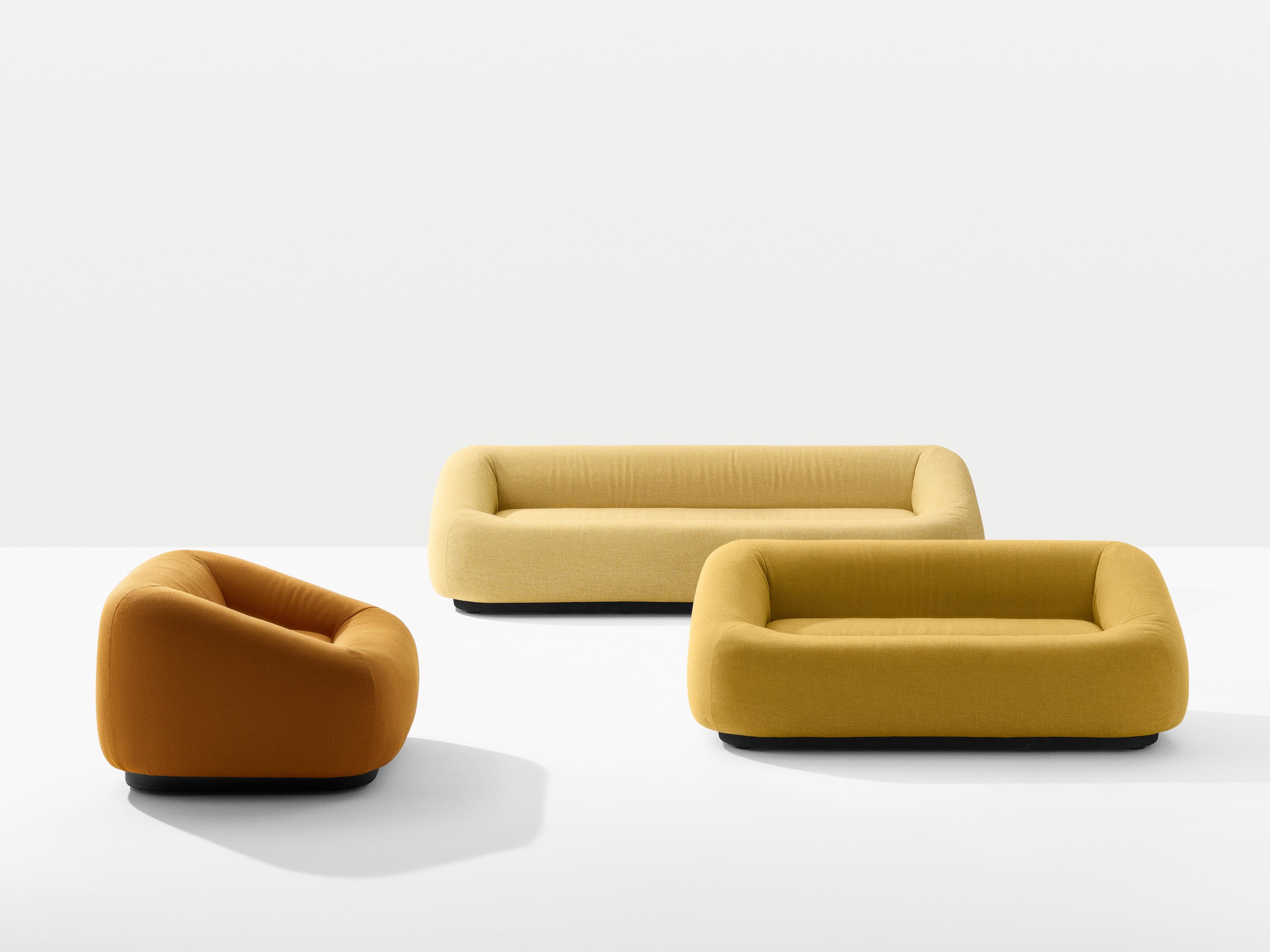 Three modern upholstered sofas in mustard yellow and orange, each with rounded edges and low black bases, are arranged on a white background.
