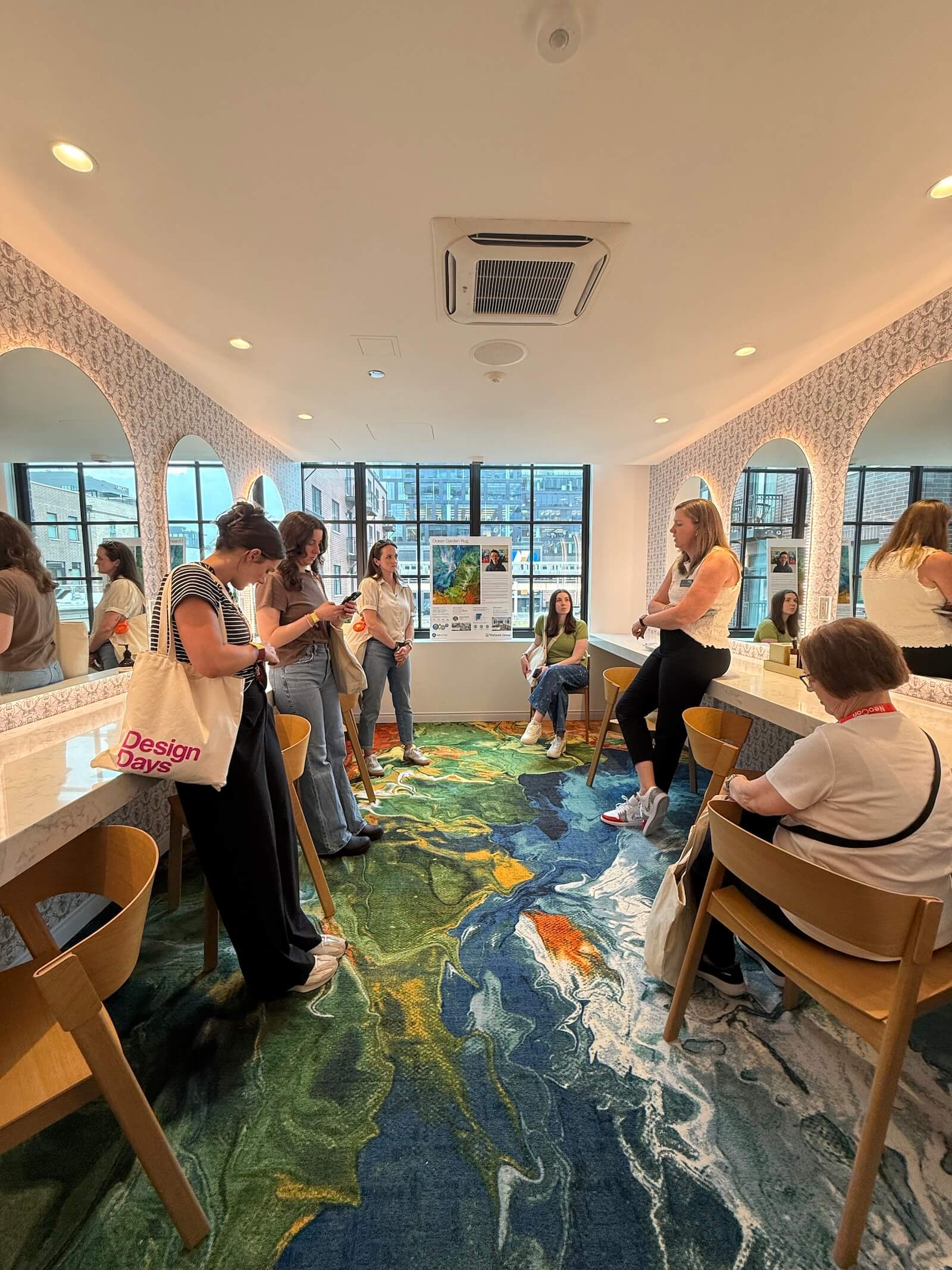 A group of people stand and sit in a modern room with mirrors, wooden chairs, and a colorful abstract carpet, some looking at their phones.