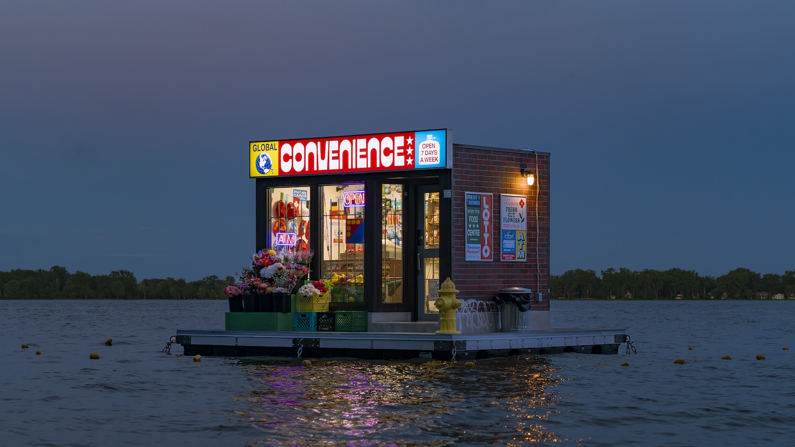 A small, brightly lit convenience store floats on a platform in the middle of a body of water at dusk, with trees visible in the background.