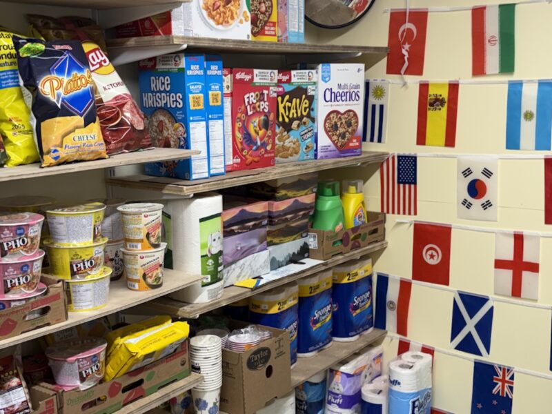 Shelves stocked with snacks, cereal boxes, and cleaning supplies; wall decorated with various international flags near a doorway.