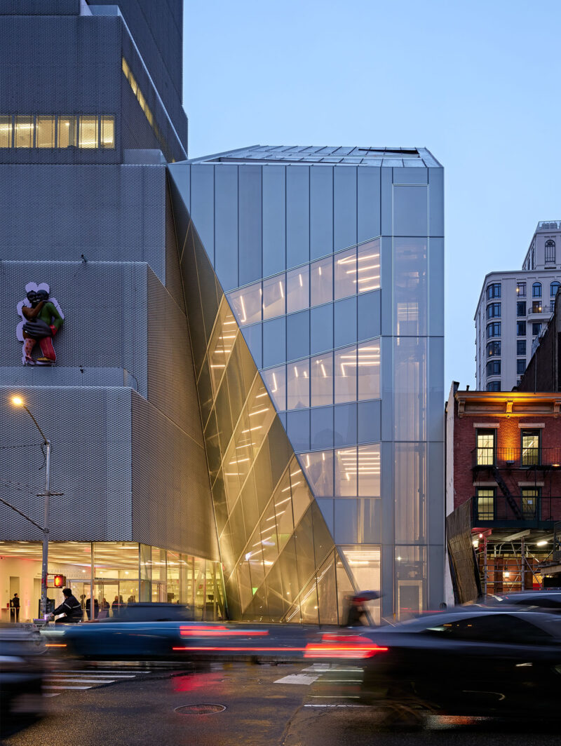 A modern glass building, home to the NYC New Museum designed by OMA, glows beside an older brick structure as traffic passes in the foreground during dusk.