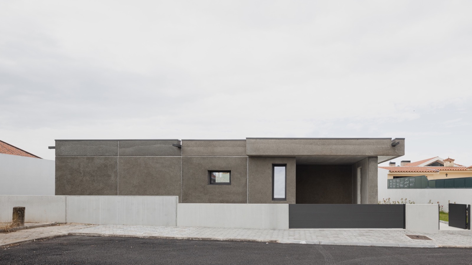 A modern, single-story house with a flat roof, minimalist gray facade, and small windows, set behind a low wall on a paved street.
