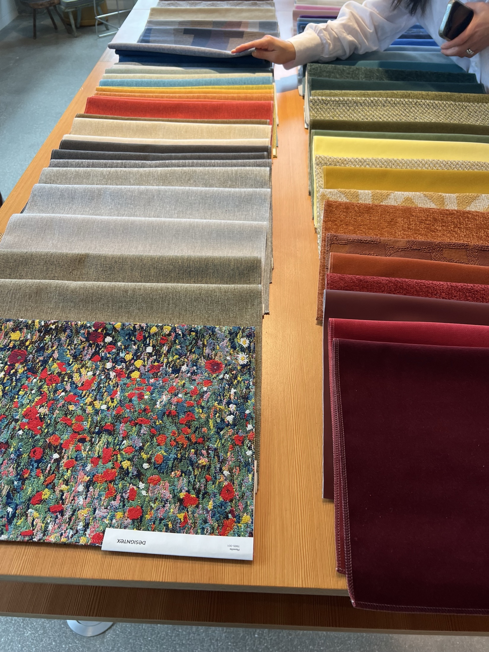 A person selects from a display of fabric swatches arranged by color on a wooden table, including patterns and solid colors.