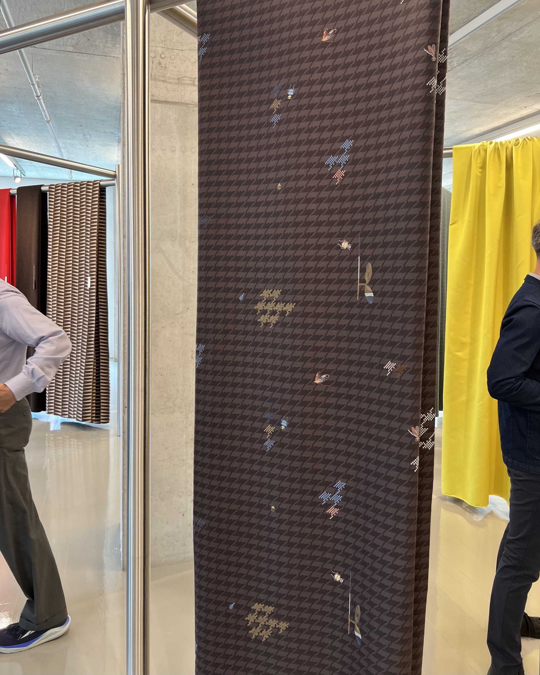 A brown patterned fabric panel with small floral and bird designs hangs vertically between metal poles, with people standing nearby and other colored panels in the background.
