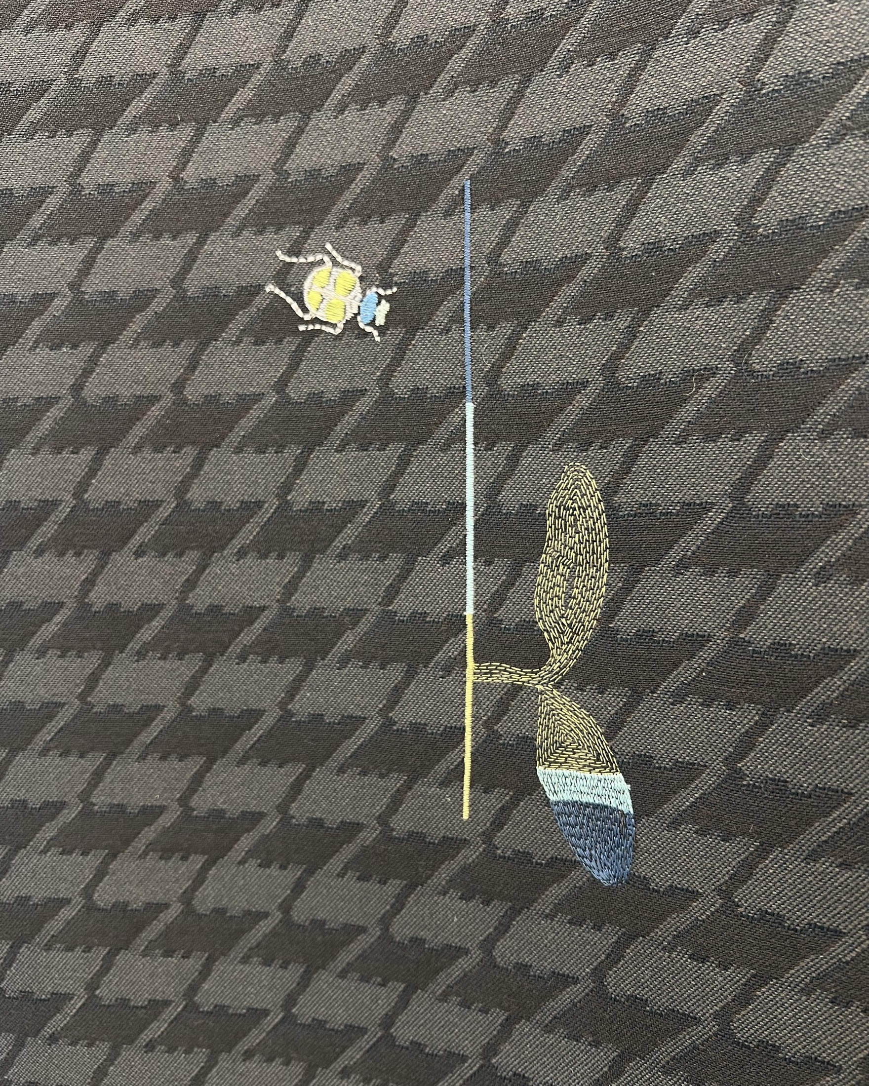 A close-up of patterned fabric with a small embroidered bee above a stylized leaf and vertical line in contrasting colors.