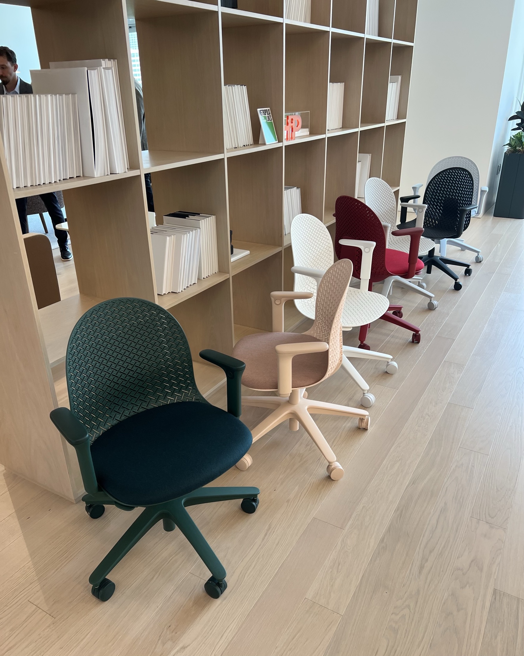Five office chairs in different colors are lined up in front of a wooden bookshelf with books and magazines in a modern office space.