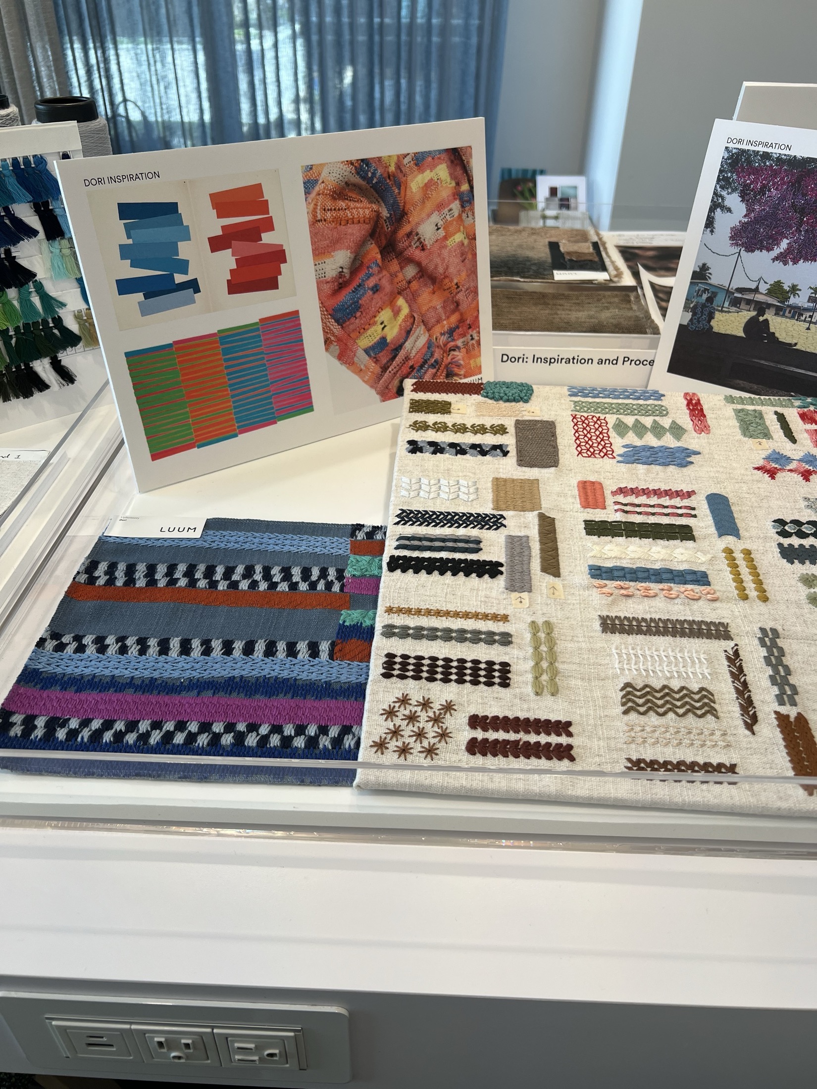 Display of textile samples and inspiration boards on a table, featuring various colorful woven and stitched patterns, with a window and curtains in the background.