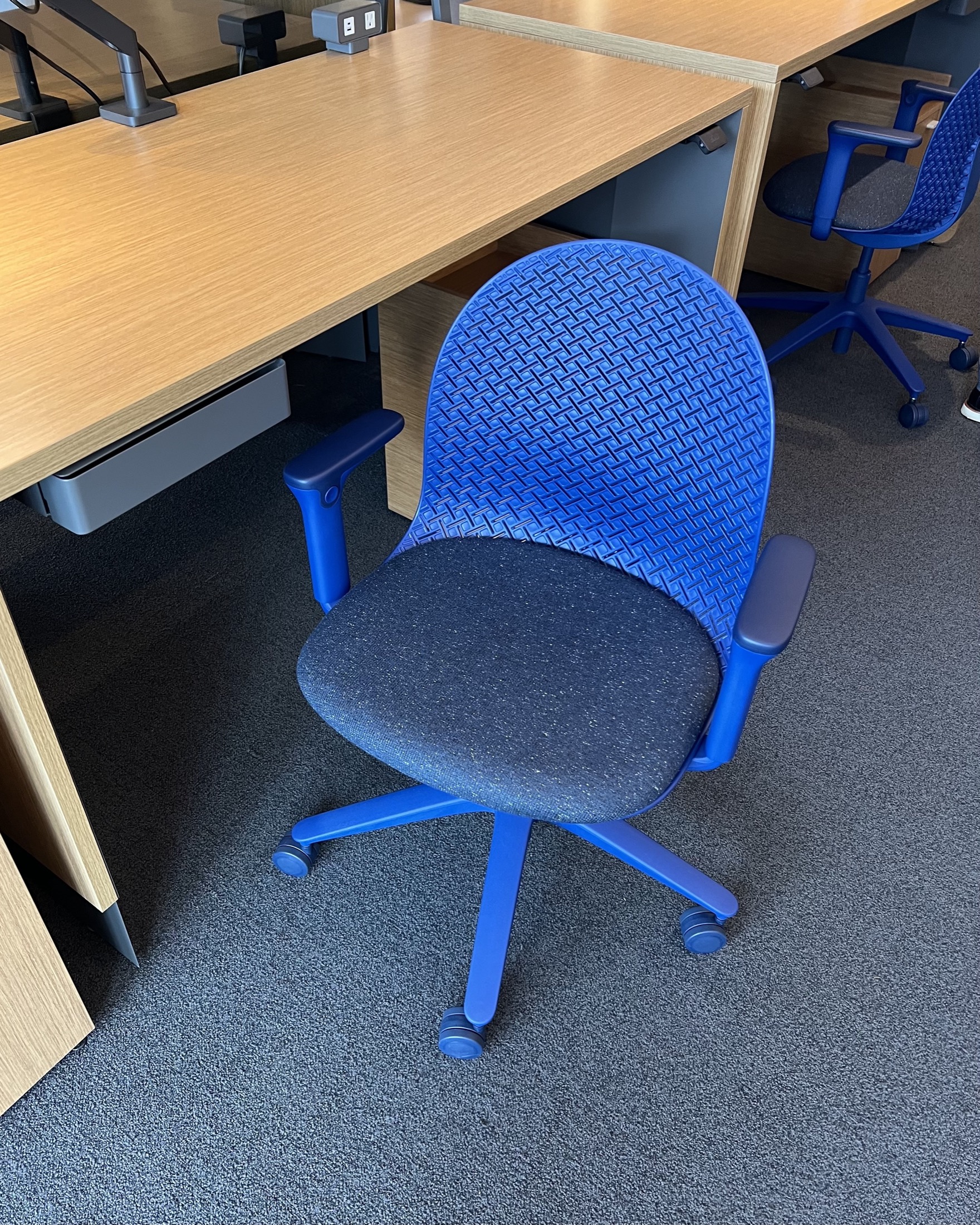 A blue office chair with armrests and wheels is positioned at a wooden desk on a carpeted floor.