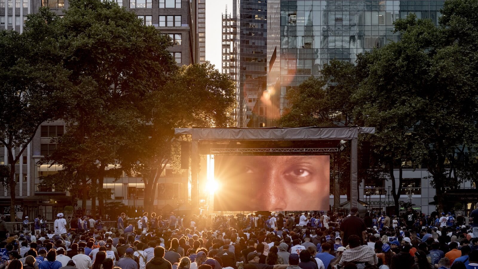 A crowd sits on grass in a city park, watching a large outdoor screen displaying a close-up of a person's eyes as the sun sets behind tall buildings.